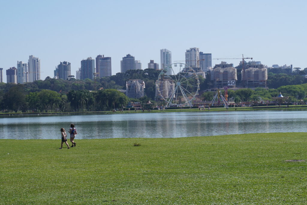 View of park Barigui with two children walking, a lake, an amusement park, and a skyline of residential building in the back.