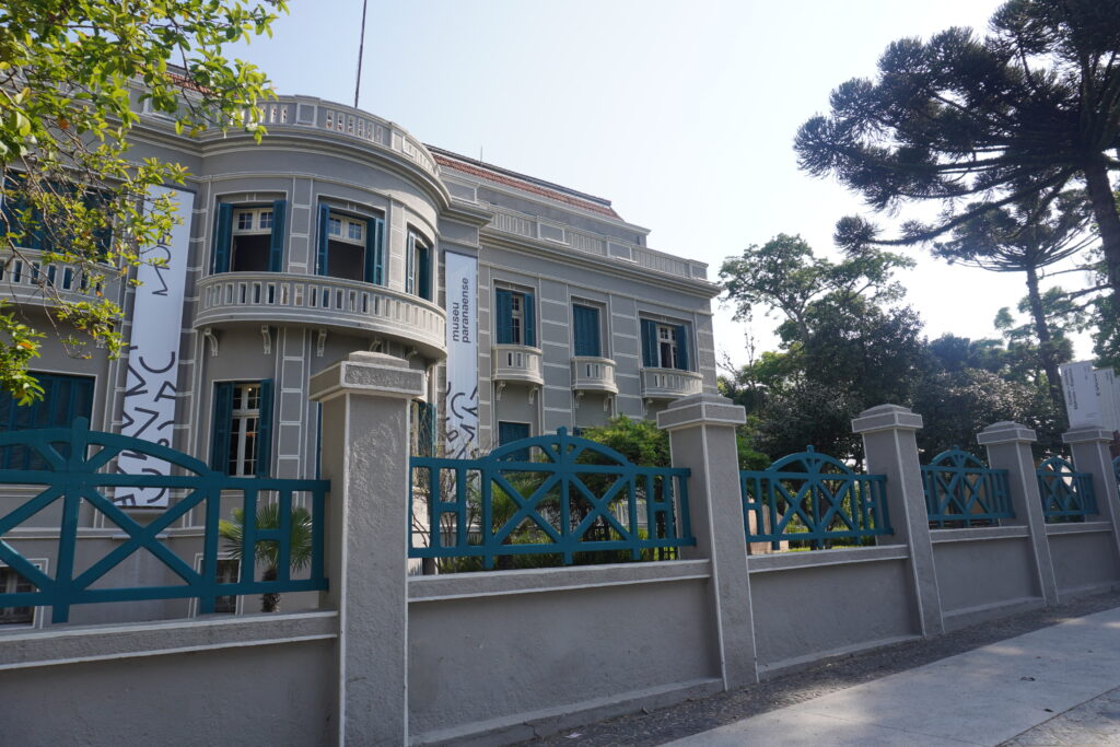 Classic-style entrance to MUPA, featuring its beige, white, and turquoise façade and araucaria trees.