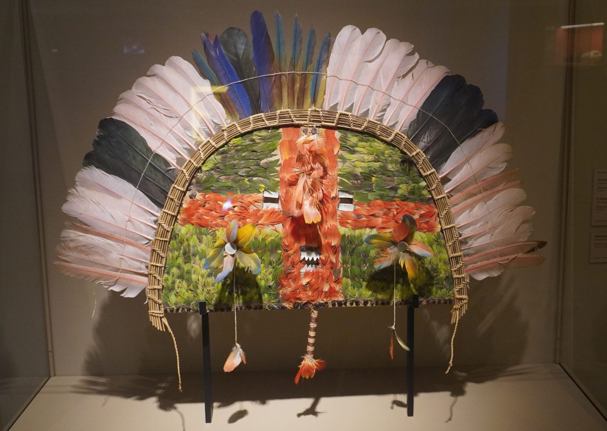 Giant piece of tribal art shaped like a half circle. It uses bird feathers to create a face, ears with earrings, and a giant crown.