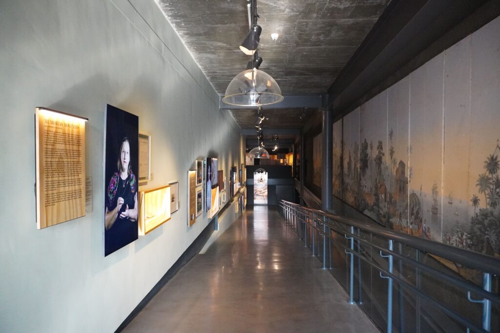 Long corridor where the walls host screens where different people talk about their immigration stories