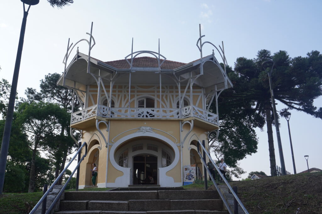 Small round-shaped yellow bulding with white finishes. Behind the building, Araucaria trees can be seen.