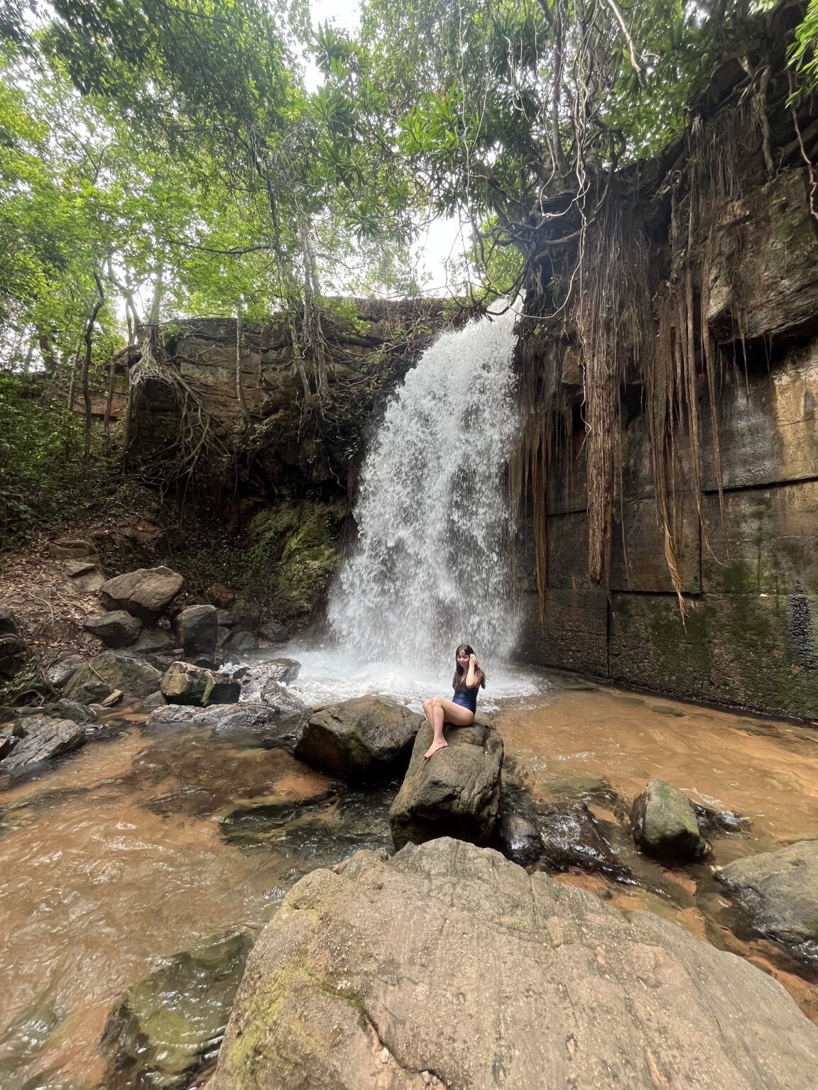 Girl sitting on stone in gront of the Salgadeira waterfall at Chapada dos Guimaraes. Human made structural reinforcement can be seen under the waterfall.