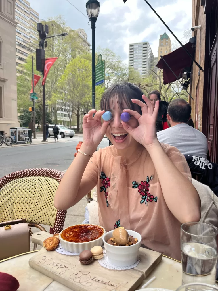 Girl sitting outdoors at Parc Restaurant in Philadelphia holding two mini macarons in front of her eyes. On the table there is lava cake, creme brulee, and more macarons!