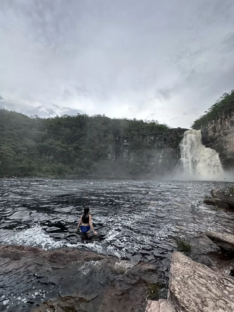 Girl walking inside the river at Chapada dos Veadeiros National Park. A maasive waterfall can be seen in the back.