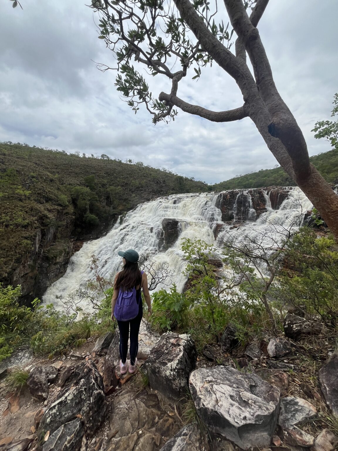 Girl wearing navy blue yoga clothes and wearing a green hat with a purple backpack standing in front of Couros Falls at Chapada dos Veadeiros