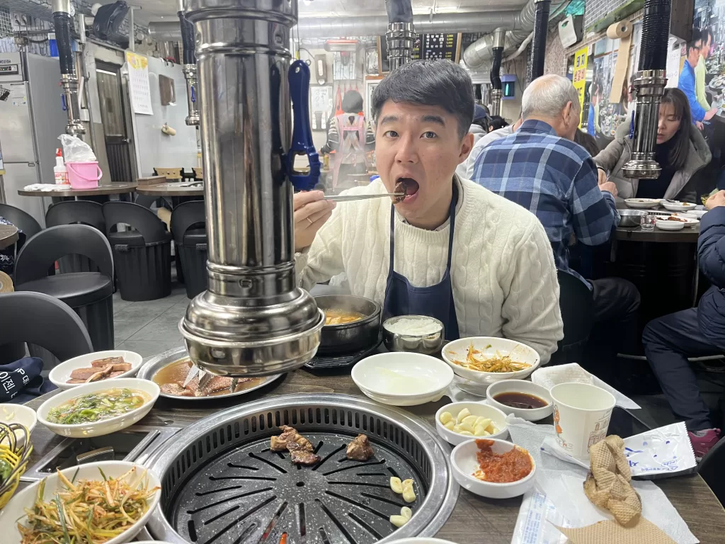 Korean man in a K-BBQ restaurant. The table includes the cooking stone and about 11 dishes with different main and side dishes.