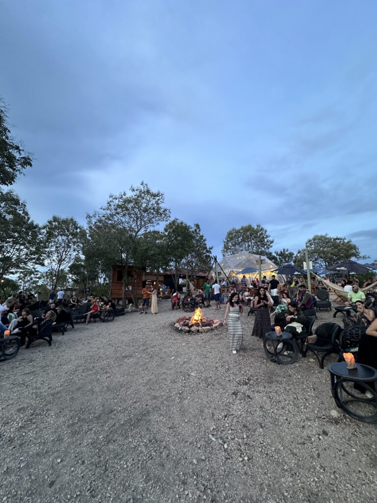 View of the sunset party with dozens of people seating in tables and hammocks. In the center there is a fireplace. In the back there is a spherical transparent structure where concerts are held.