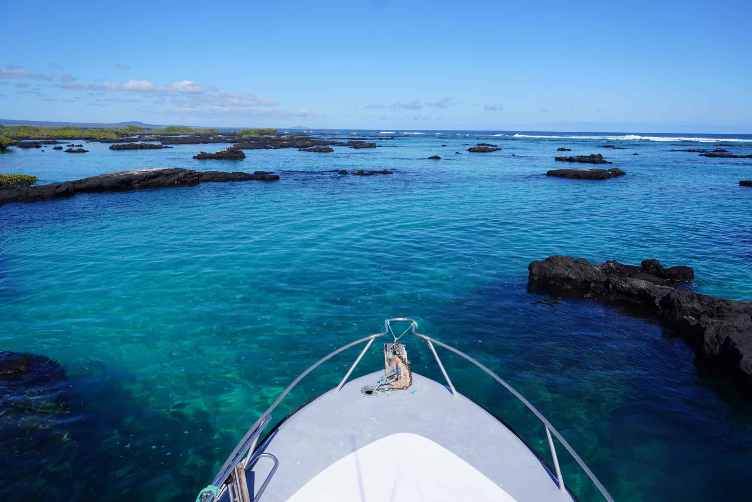 View of the front of a motorboat looking into the open ocean. Waters are turquoise and blue, and there are lots of dots of black vulcanic stones in the horizon.