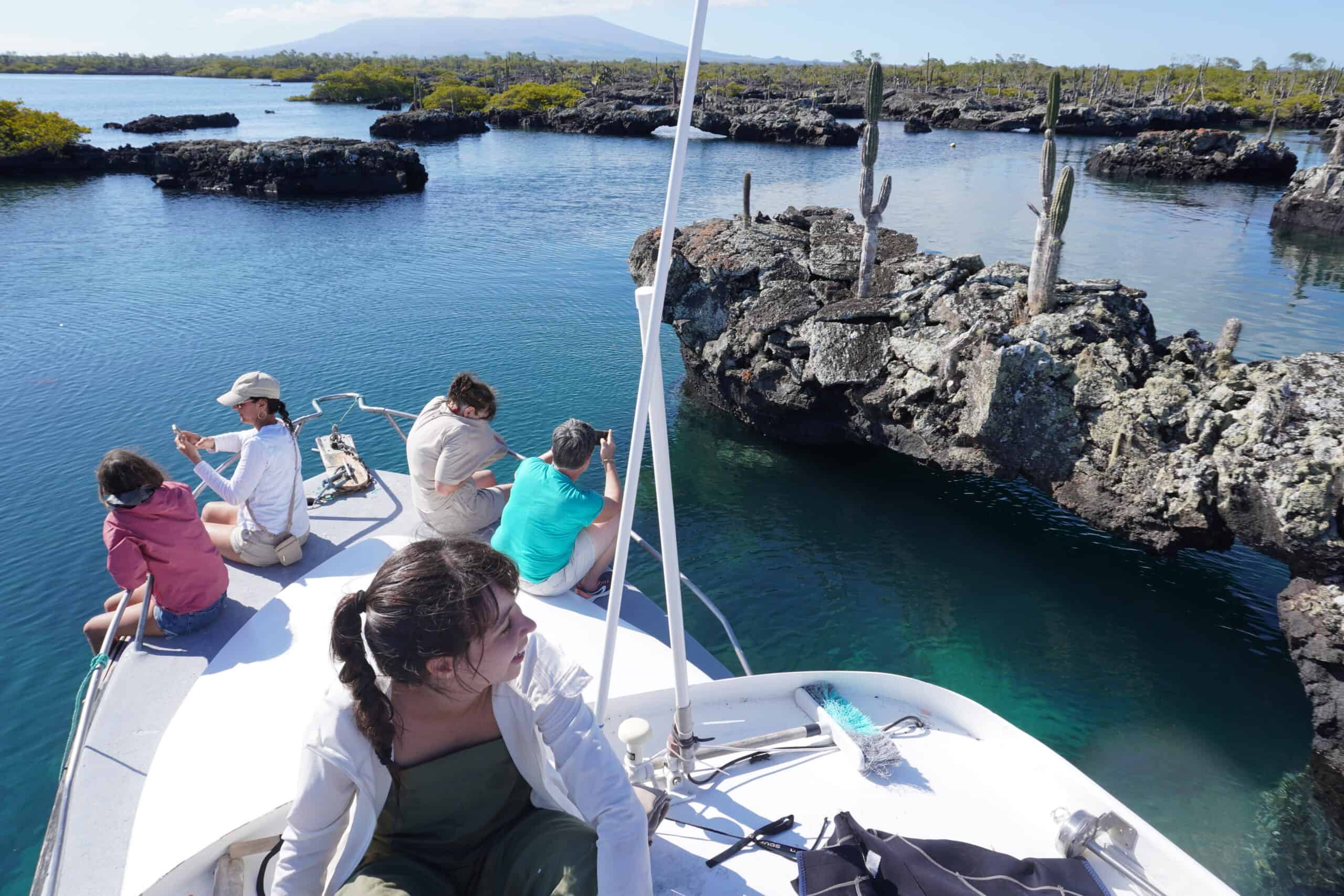 5 people sitting on a motorboat and enjoying the Los Tuneles scenery. The landscape contains volcanic rocks forming arch shapes or small round islands. Long and tall cactuses grow from the rocks.