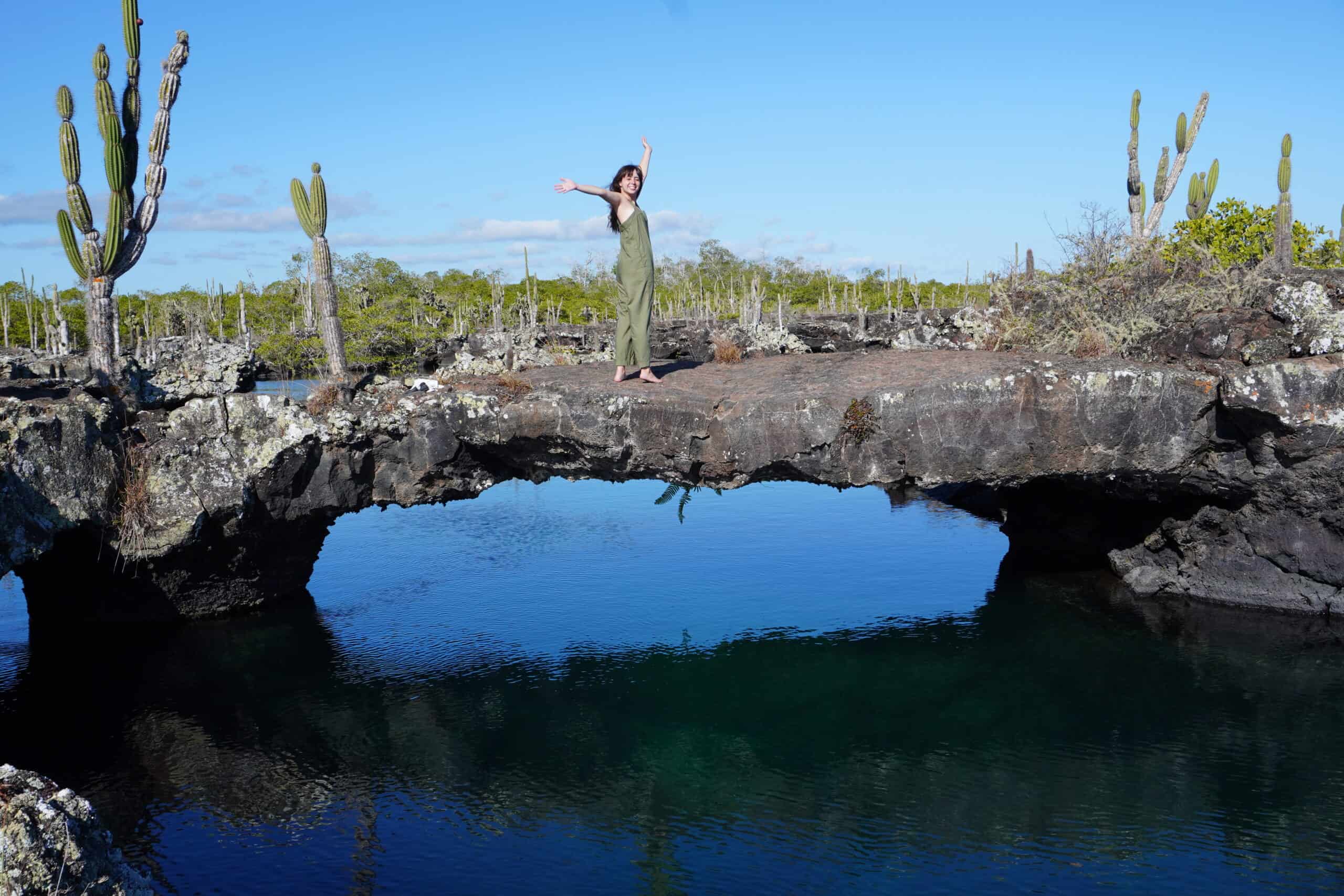 Lo Lee standing in a volcanic rock arch above the ocean. Cactuses grow on the sides. On the back mangrove and the Los Tuneles landscape can be seen.