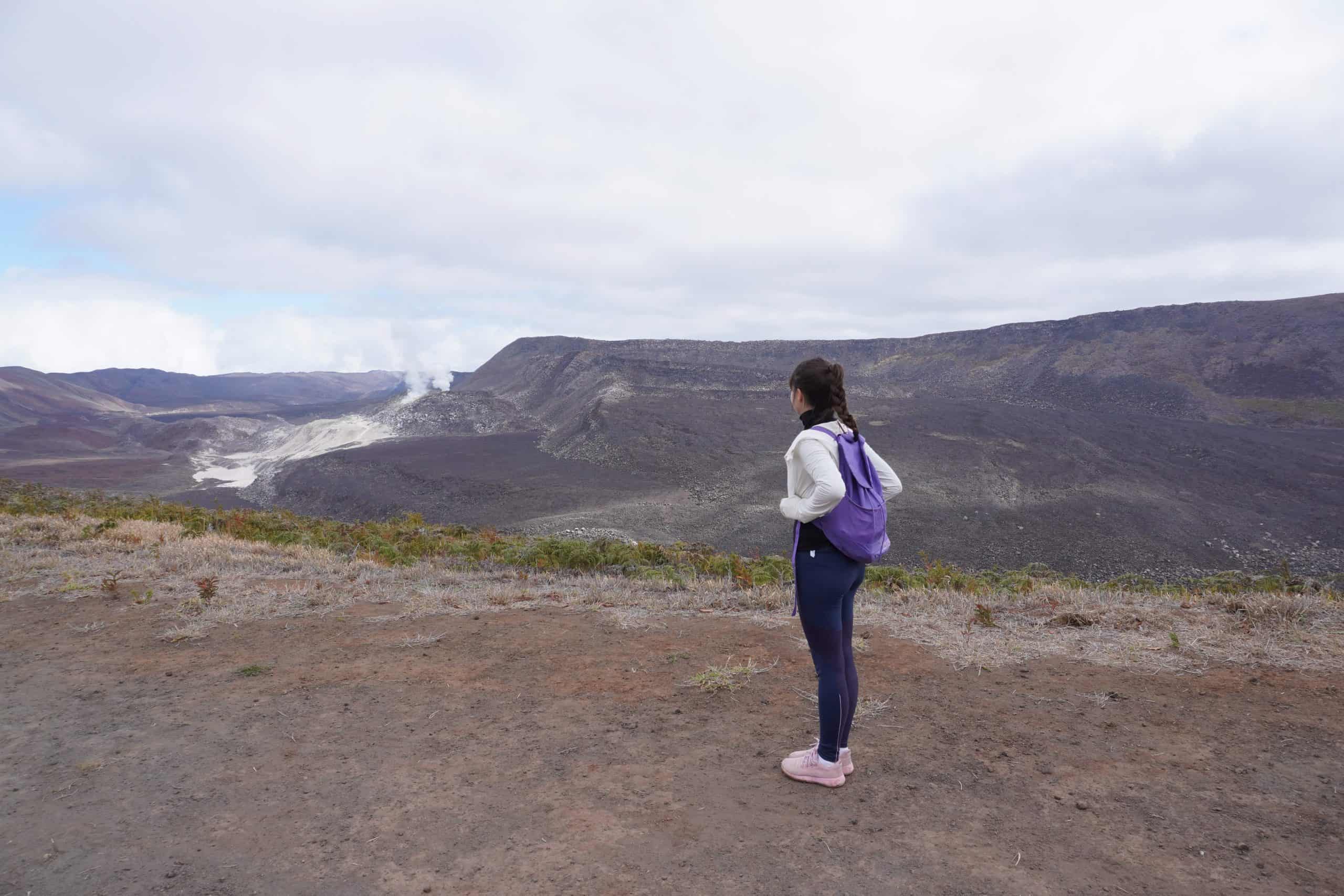 Girl wearing hiking clothes stands on the road leading to the trail at Sierra Negra Volcano. The day is cloudy and the sulfur mines of Sierra Negraa volcanos can still be seen from a distance.