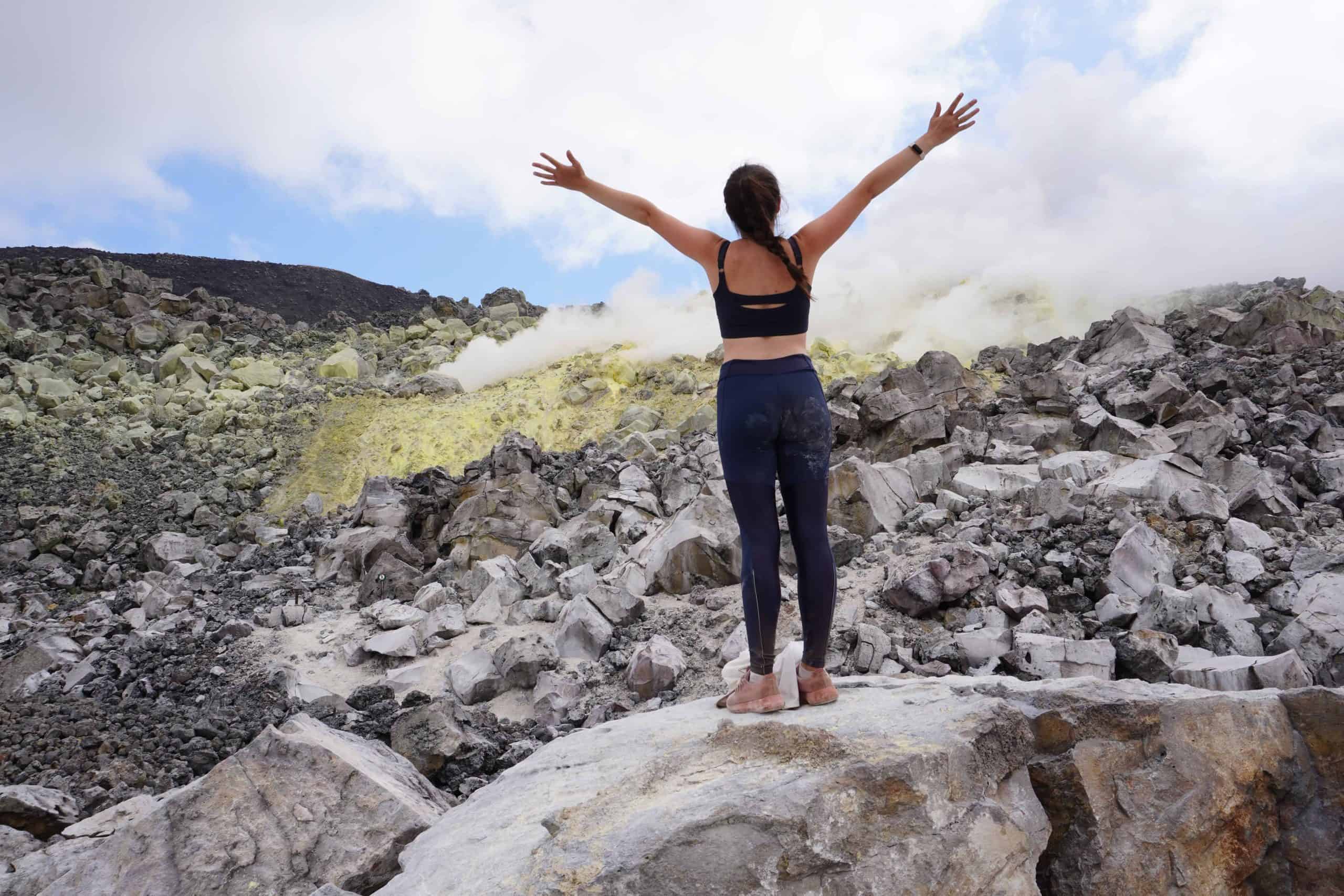 Girl standing on rock with her arms open while looking at the yellow and smoking sulfur mines in the Sierra Negra Volcano Hiking Tour. She is standing only 10 meters away from the active sulfur vents.