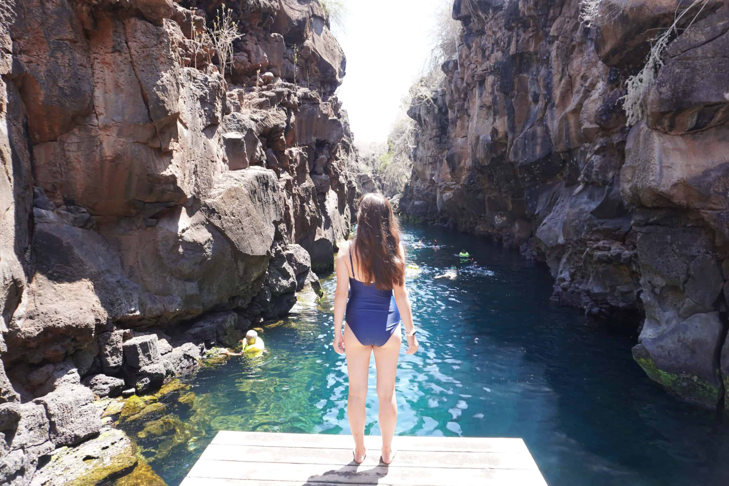 Girl standing in front of the water in Las Grietas, Galapagos. The water is bright blue and many snorkelers swim slong the channel.