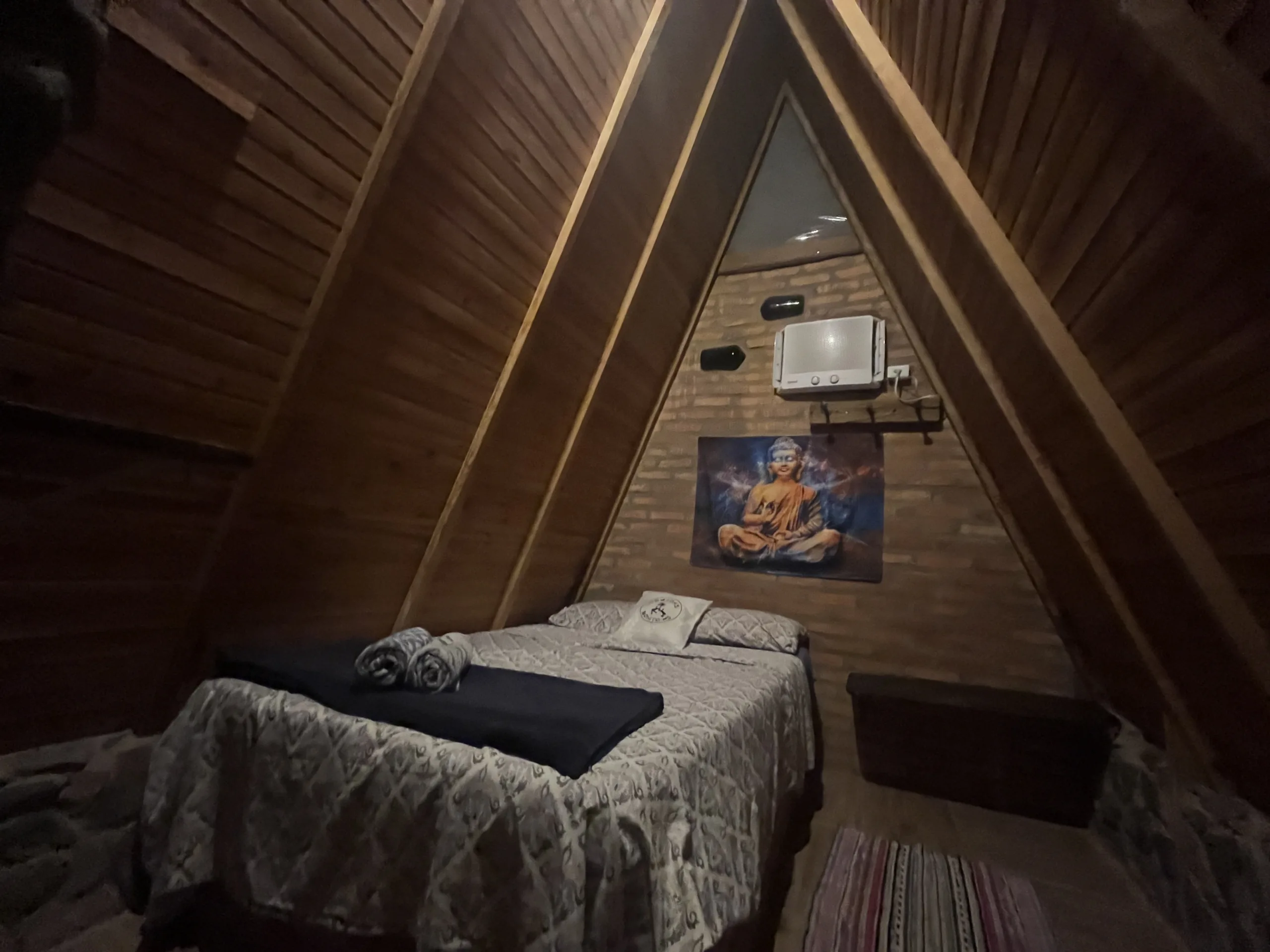 Inside of cabin at Nomadas Bonito. The cabin is triangular and made of wood. There is one bed, air conditioning, and a tapestry of Buddha