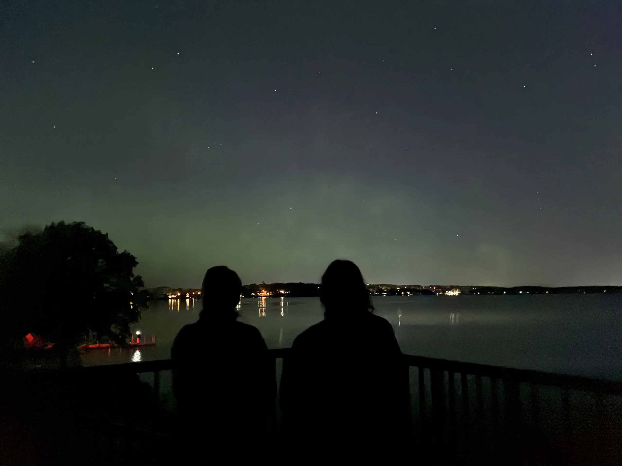 Two girls looking over the lake at Marshall park to enjoy a view of green nothern lights. City lights can be seen in the horizon