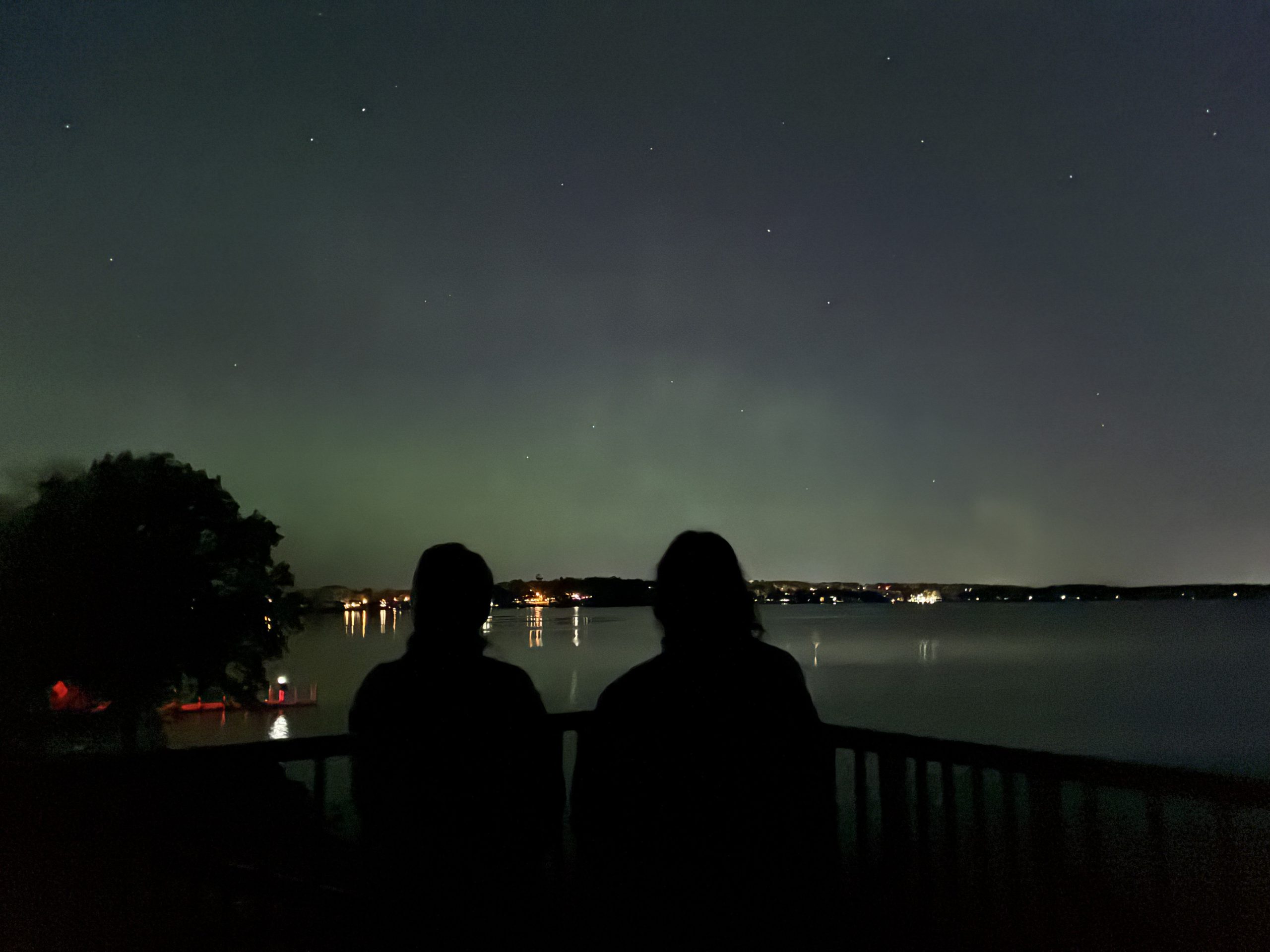 Two girls looking over the lake at Marshall park to enjoy a view of green nothern lights. City lights can be seen in the horizon