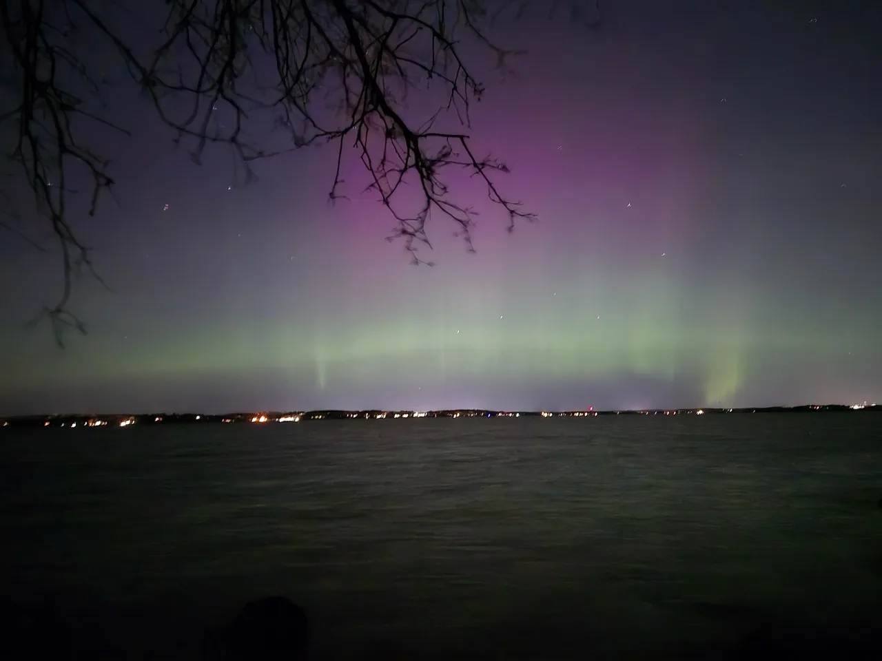 Northern lights shining bright above the lake at Raymer's Cove in Madison, Wisconsin. The Aurora is bright, pink on the top and green on the bottom. In the horizon, the city lights can be seen.