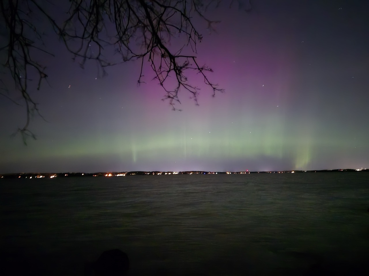 Northern lights shining bright above the lake at Raymer's Cove in Madison, Wisconsin. The Aurora is bright, pink on the top and green on the bottom. In the horizon, the city lights can be seen.