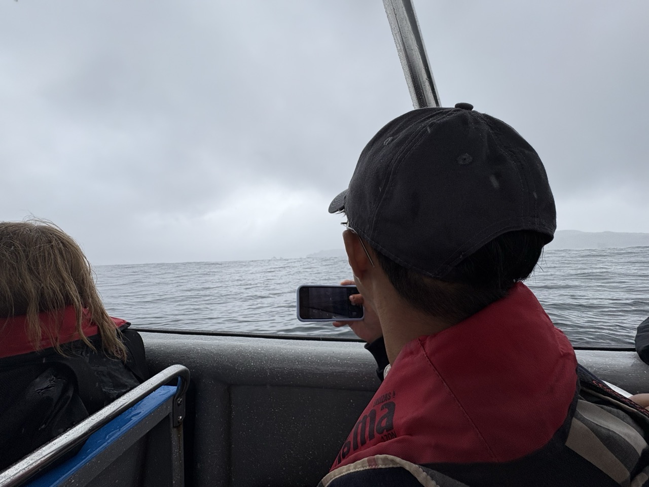 Man sitting in a small motor boat during the Isla Chanaral whale watching tour. He is pointing his phone towards the ocean in the hopes of photographing a whale.